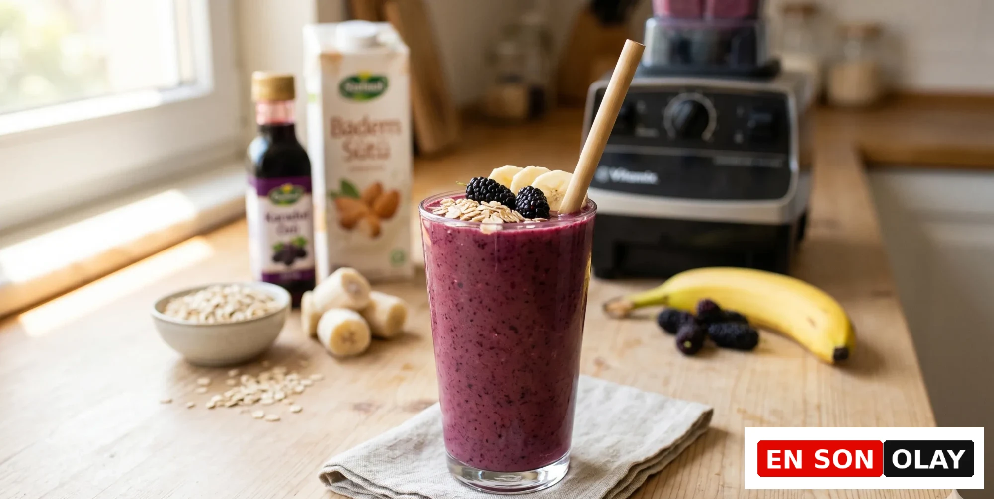 Ahşap tezgah üzerinde duran, karadut özlü mor renkli bir sağlıklı smoothie, yulaf ezmesi, muz dilimleri ve taze karadutlarla süslenmiş bir cam bardakta, arka planda badem sütü ve karadut özü şişesiyle beraber Vitamix blenderın da görüldüğü bu tarif için ferahlatıcı bir görüntü sunuyor.