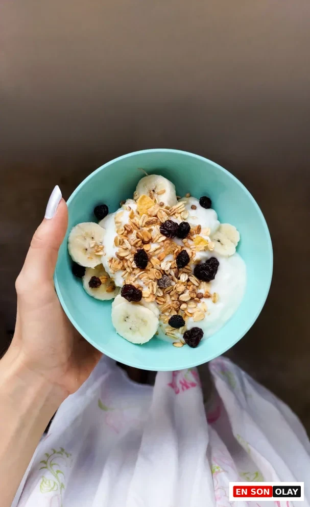 Beyaz ojeli bir elin tuttuğu, içinde dilimlenmiş muz, granola ve kuru üzüm ile süslenmiş açık mavi bir kase yoğurt, sağlıklı bir probiyotik kaynağı olarak fotoğrafın merkezinde yer alıyor.