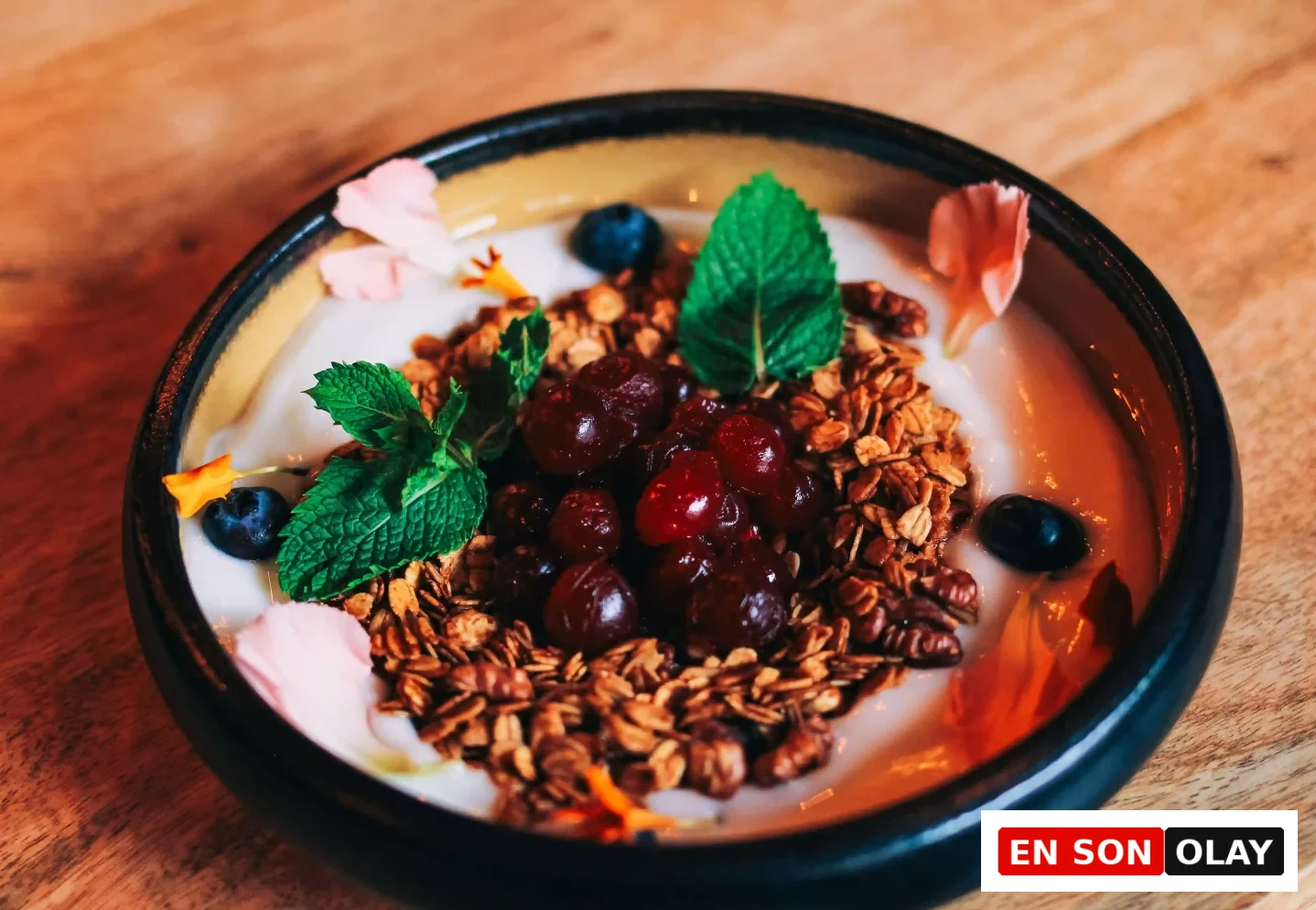 Koyu renkli bir kasenin içinde, probiyotik zengini, taze yoğurt üzerine serpiştirilmiş kıtır granola, koyu kırmızı taneli meyveler, parlak mavi yaban mersinleri, taze nane yaprakları ve renkli yenilebilir çiçek yapraklarıyla süslenmiş, sağlıklı ve göz alıcı bir tabak.