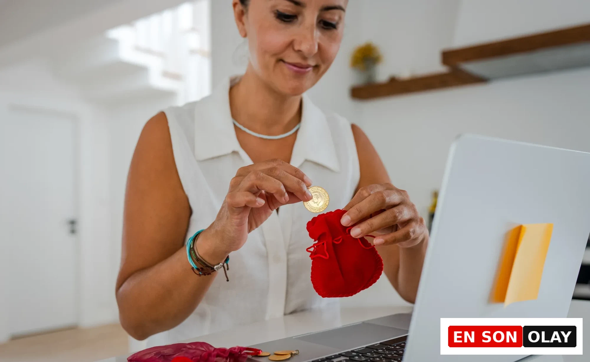 Koç burcu kadını, nisan ayında beklenen finansal kazanç kapılarını simgelercesine elinde altın para tutarak kırmızı kadife keseye özenle yerleştirirken, önündeki açık laptop ile finansal işlemlerini yönetircesine gülümsüyor.