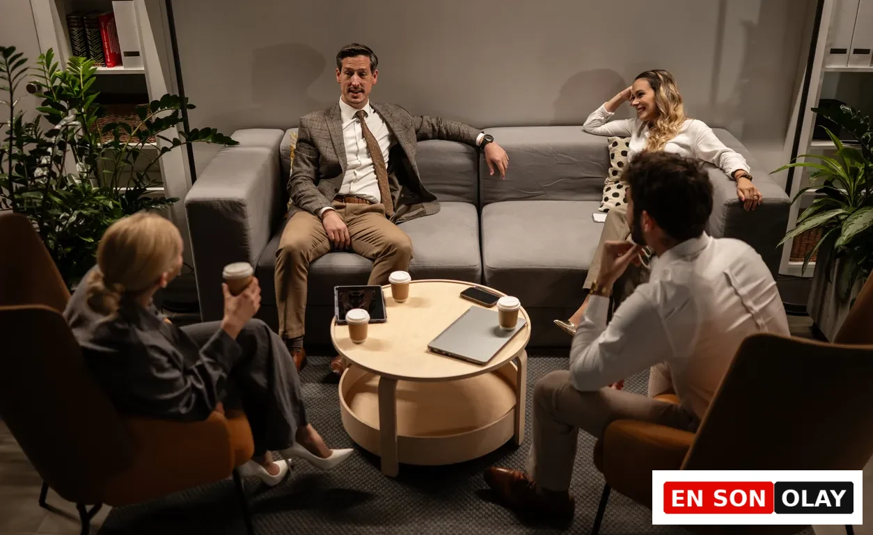 Modern bir ofis lounge alanında, gri bir kanepe ve iki kahverengi koltukta oturan dört iş arkadaşı, yuvarlak ahşap sehpada duran kahveler, tablet, akıllı telefon ve laptop eşliğinde samimi bir iş toplantısı gerçekleştiriyor, bu görüntü iş hayatındaki verimli ekip toplantılarının rahat bir ortamda da yapılabileceğini vurguluyor.