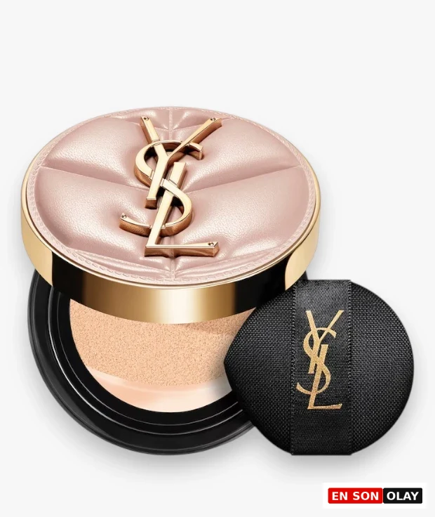 Yves Saint Laurent Skin Affair Cushion Fondöten, kapitone desenli açık pembe kapağında altın rengi YSL logosu taşıyan yuvarlak şık bir kutuda, açık renkli fondöten süngeri görünecek şekilde açılmış ve yanında altın YSL logosu işli siyah makyaj süngeriyle birlikte zarifçe duruyor.