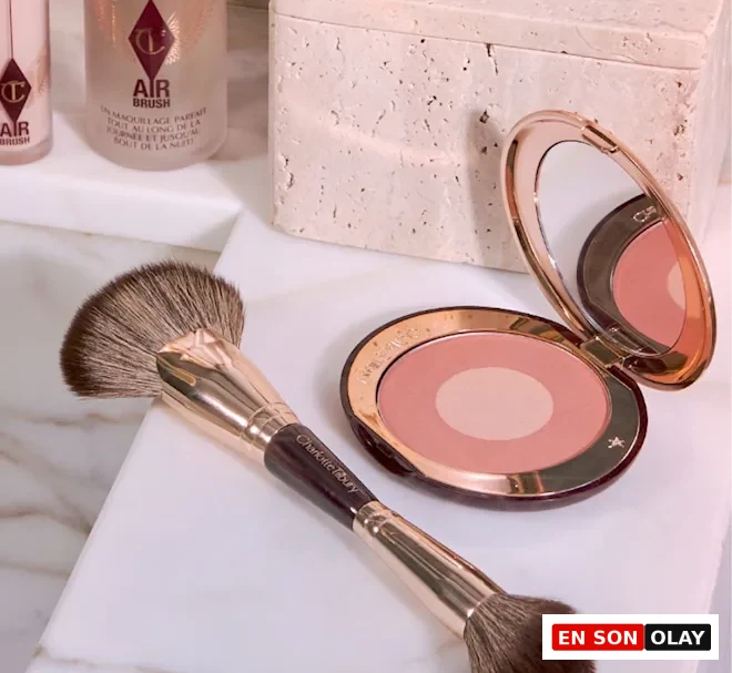 Mermer bir yüzeyde, altın renkli detaylara sahip, farklı uygulamalar için iki ucu bulunan Charlotte Tilbury makyaj fırçası, ortası açık pembe, dışı koyu pembe tonlarında allık içeren ve aynası açık duran kompakt bir paletle birlikte gösterilirken, arka planda Airbrush serisinden fondöten şişeleri de seçilebiliyor.