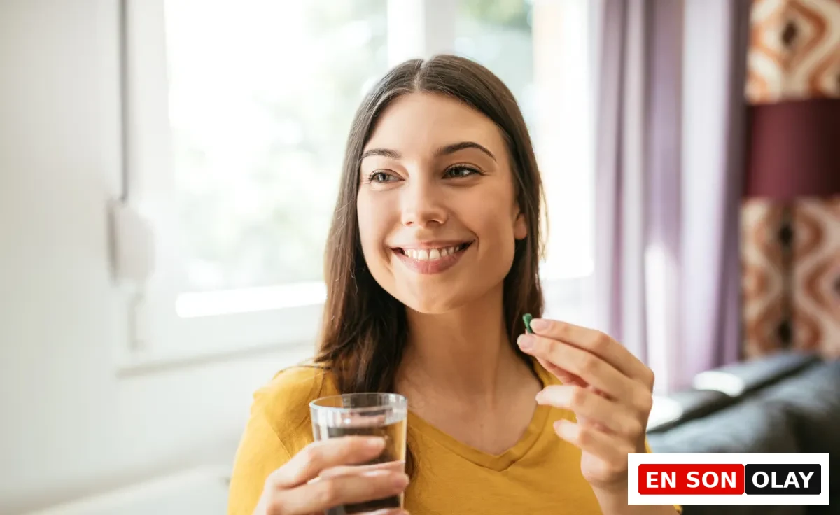 Pencereden gelen doğal ışıkla aydınlanmış bir iç mekanda, hardal sarısı bir bluz giymiş gülümseyen genç bir kadın, L-theanine takviyesi kullanımını gösterir şekilde, sol elinde berrak bir bardak su tutarken sağ elinde küçük yeşil bir kapsülü ağzına götürüyor.