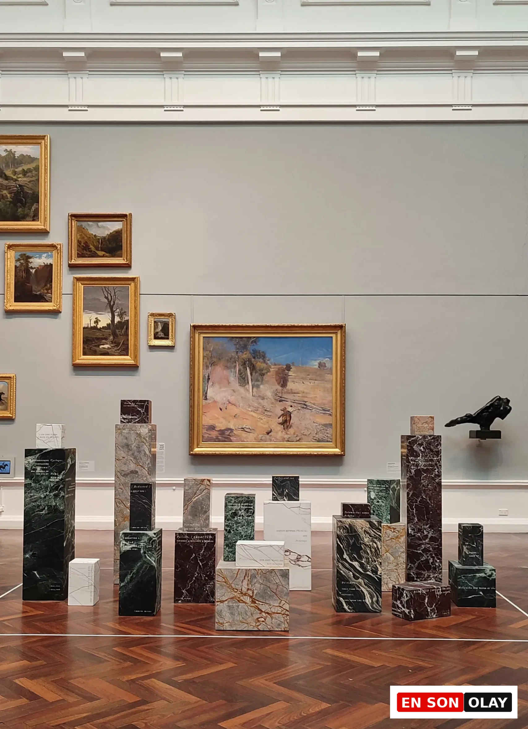 Sanat galerisinde, ahşap parke zemin üzerinde yükselen, üzerlerinde yazılar bulunan çeşitli renk ve desenlerdeki mermer bloklardan oluşan ilgi çekici bir modern sanat enstalasyonu, arkada duvarda asılı geleneksel yağlıboya manzara resimleriyle beraber sergileniyor.