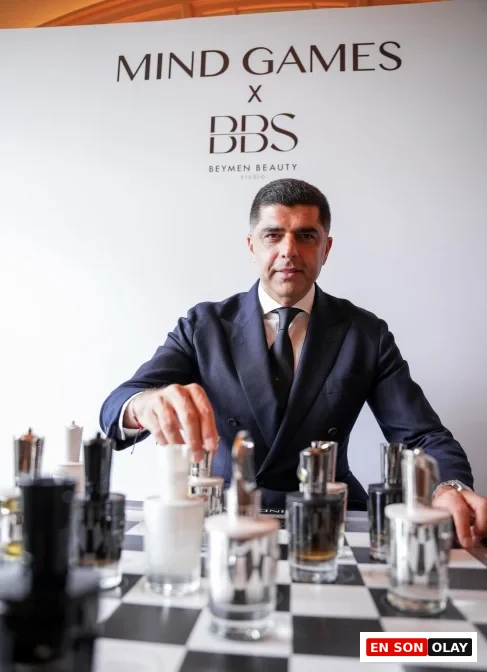 Mind Games Parfum kurucusu Alex Shalbaf, bir röportaj sırasında koyu renk takım elbisesiyle oturmuş, ellerini birleştirmiş bir şekilde hafifçe gülümserken profesyonel ve düşünceli bir duruş sergiliyor.