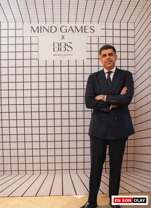 Mind Games Parfüm kurucusu Alex Shalbaf, şık bir takım elbiseyle oturduğu, arkasında modern iç mekan detaylarının bulanıkça seçildiği, röportaj anından yakalanmış, düşünceli ifadesiyle objektife bakan profesyonel bir portrede yer alıyor.