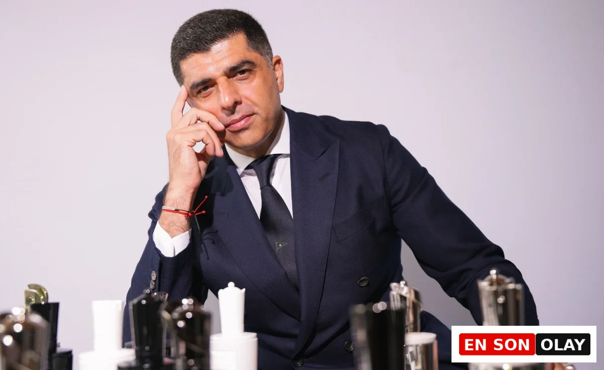 Mind Games Parfüm kurucusu Alex Shalbaf, zarif koyu renk takım elbisesi içinde, özenle tasarlanmış Mind Games parfüm şişelerinin masada belirgin bir şekilde durduğu bir ortamda, kameraya doğrudan bakarak markasının felsefesini ve estetiğini yansıtan düşünceli bir ifade sergiliyor.