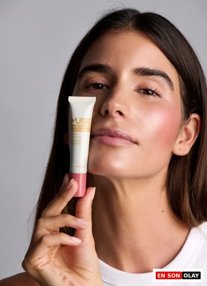 Yanaklarına uyguladığı doğal kiraz krem allık ile sağlıklı bir ışıltıya sahip, uzun koyu saçlı bir kadın, beyaz bir tişört giymiş ve elinde üzerinde Muse Lab markalı Cheek to Cheek Cherry Cream Blush yazan küçük bir allık tüpü tutuyor.