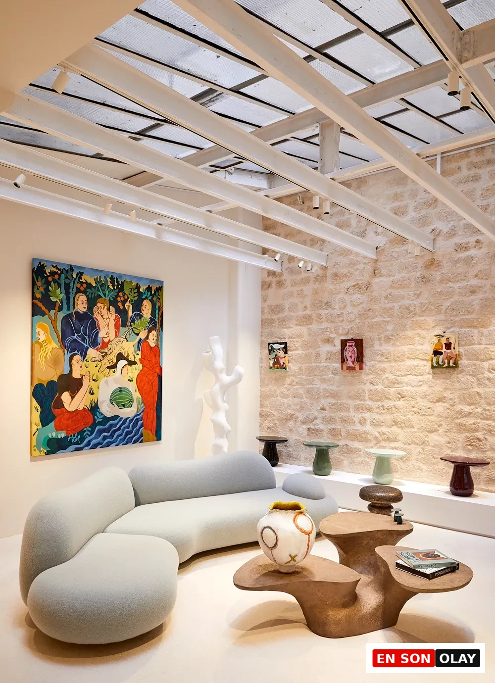 Paris Tasarım Haftası kapsamında sergilenen modern bir iç mekan düzenlemesinde, geniş renkli bir tablonun önünde uzanan kıvrımlı, açık gri bir koltuk takımı, sanatsal bir vazo ve bir kitabın eşlik ettiği özgün tasarımlı organik formlu bir sehpa dikkat çekerken, taş duvar üzerindeki minik tablolar ve çeşitli renklerdeki yuvarlak masalar çağdaş tasarımın estetiğini gözler önüne seriyor.