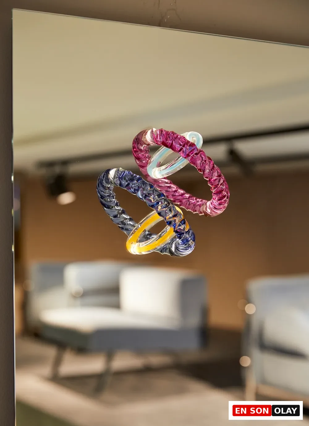 Bu üç boyutlu, pembe, mavi ve şeffaf cam halkalardan oluşan sanat eseri, Paris Tasarım Haftası'nın ruhunu yansıtan modern bir tasarım ve sanat enstalasyonu olarak bir aynanın önünde etkileyici bir şekilde sergileniyor; aynada bulanık iç mekan yansıması görülüyor.