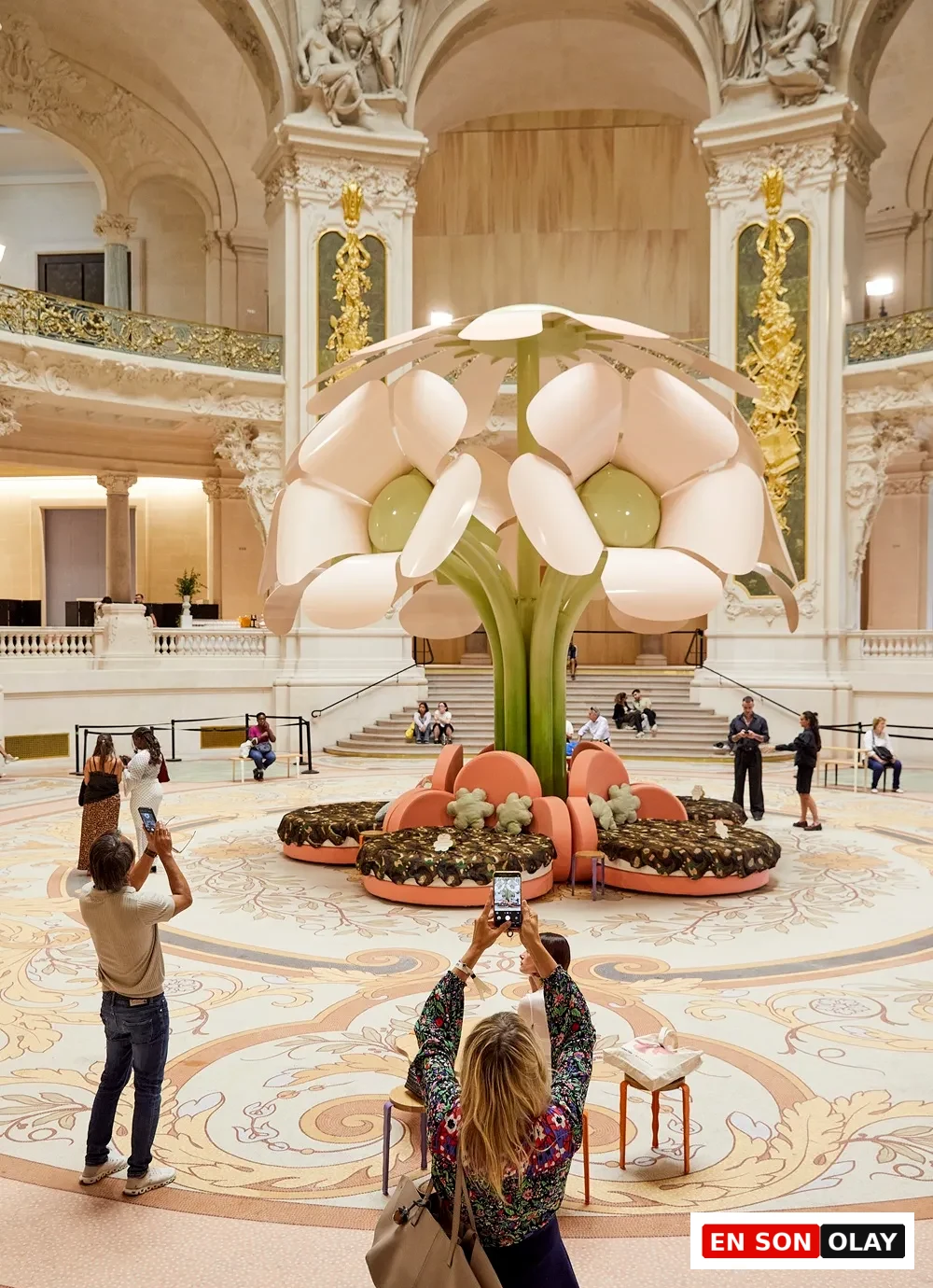 Geniş, yüksek tavanlı ve altın işlemeli detaylara sahip gösterişli bir salonda, ortada Uchronia'nın Paris Design Week için tasarladığı, açık pembe yaprakları ve yeşil gövdesi olan devasa bir çiçek formunda enstalasyon duruyor, etrafında desenli zeminde fotoğraf çeken ve dinlenen ziyaretçilerle birlikte çağdaş bir tasarım sergisini yansıtıyor.