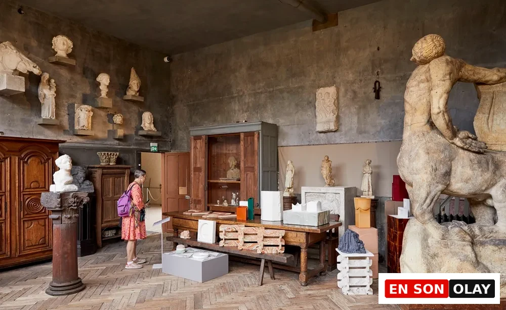 Bourdelle Müzesi'nin parke zeminli, koyu renkli duvarlara sahip tarihi bir salonunda, klasik heykellerin ve büyük sentor heykelinin ortasında Paris Tasarım Haftası için sergilenen modern tasarım objelerini dikkatle inceleyen bir ziyaretçi.