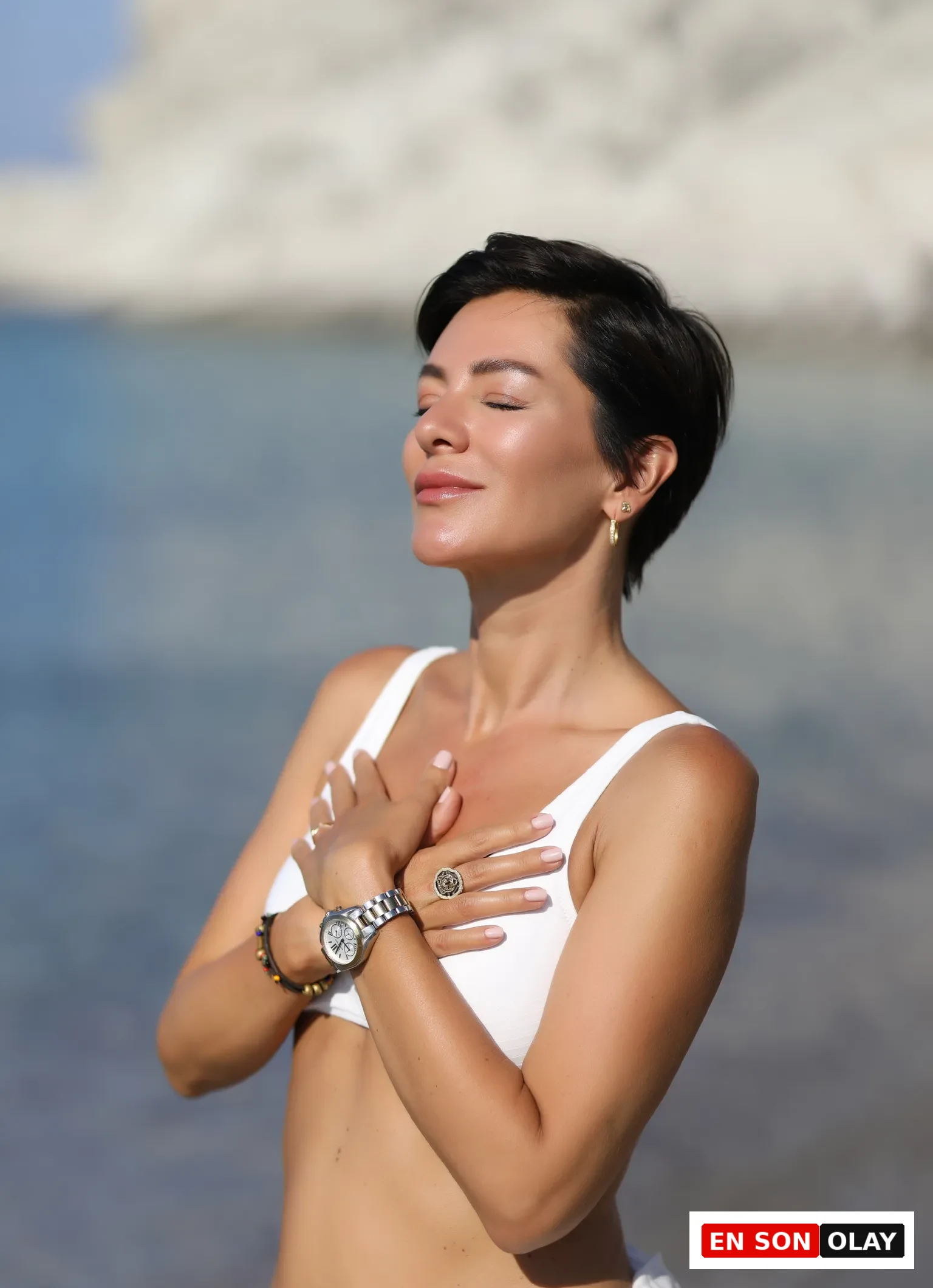 Güneşli bir günde, kısa siyah saçlı, gözleri kapalı ve huzurlu bir ifadeye sahip genç bir kadın, beyaz bikini üstü ve altına giydiği beyaz bir parça ile ellerini kalbinin üzerinde kavuşturmuş, arkasında mavi denizin ve beyaz kayalıkların uzandığı doğal bir ortamda duruyor.