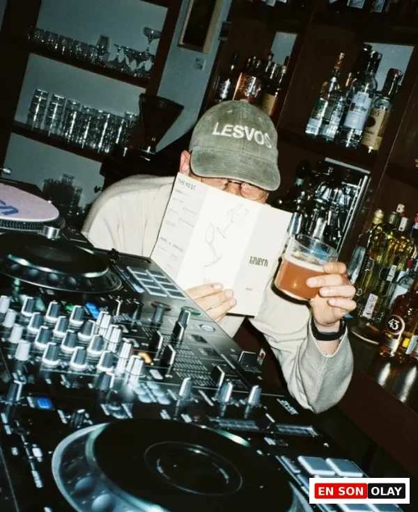 Ön planda bir DJ mikseri bulunurken, yeşil LESVOS şapkalı bir kişi elinde bir menü ve bira bardağı tutarak bar tezgahının arkasında, şişelerin ve bardakların olduğu loş ışıklı bir ortamda müzik yapmaya hazırlanıyor veya ara veriyor gibi görünüyor.