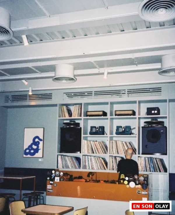 Beyoğlu'ndaki bir taproom'un iç mekanında, geniş bir plak koleksiyonu ve DJ ekipmanlarıyla dolu rafların önünde sırtı dönük bir DJ'in müzik yaptığı, turuncu bir bar tezgahı ve şık oturma alanlarının bulunduğu canlı bir atmosfer yansıyor.