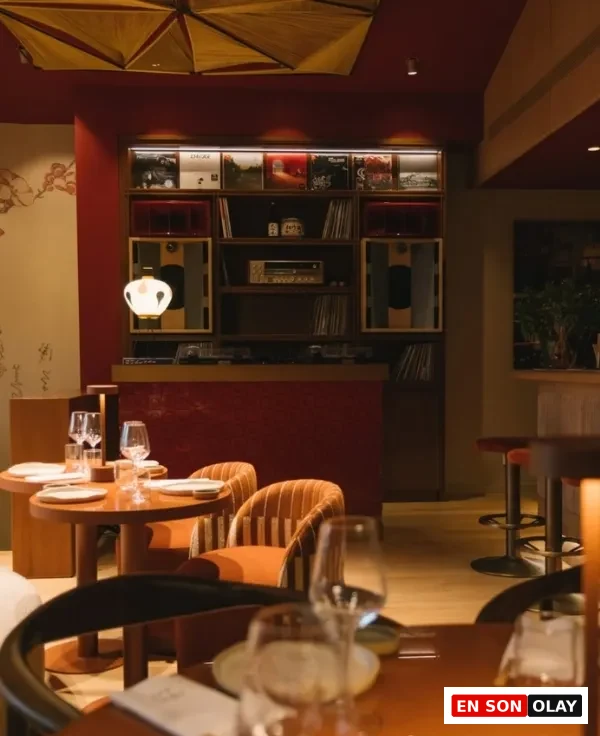 Koyu kırmızı duvarların önünde, plaklar ve ses sistemleriyle dolu özel bir nişin de bulunduğu, sıcak ışıklandırılmış modern restoranda, şık bir şekilde düzenlenmiş yemek masaları ve kadife görünümlü turuncu sandalyeler dikkat çekiyor.