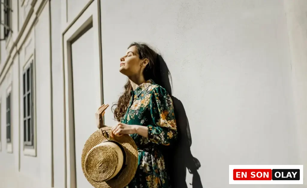 Güneş ışığında yüzünü yukarı kaldırarak duvara yaslanan, elinde hasır şapka tutan çiçek desenli elbise giymiş kadın