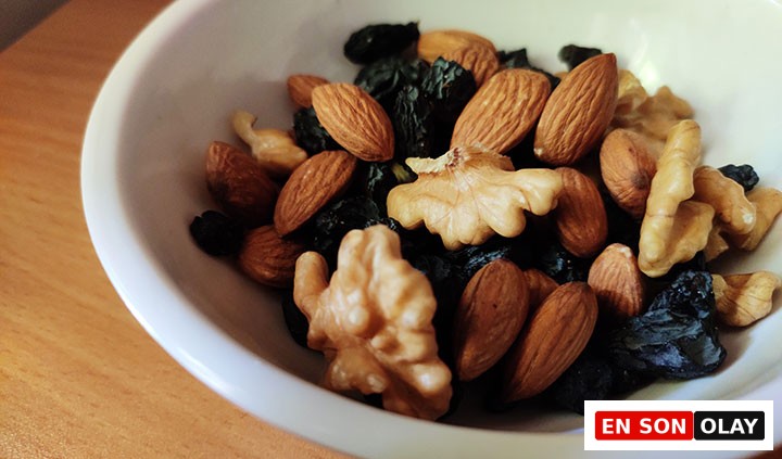 Ahşap bir masa üzerinde, parlak beyaz bir kase içerisinde, saç derisi sağlığını destekleyen ve Omega-3 gibi faydalı yağlar içeren badem, ceviz ve koyu renkli kuru üzüm tanelerinden oluşan besleyici bir karışım yakın planda yer alıyor.