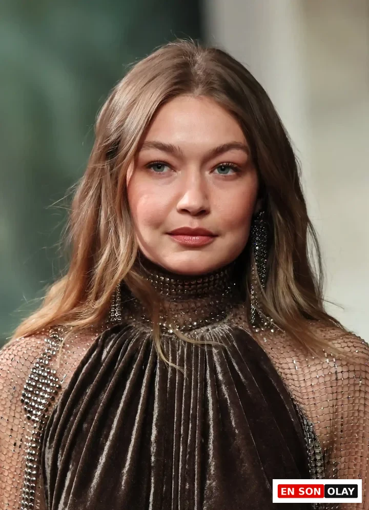 Gigi Hadid, Ralph Lauren podyumunda, ışıltılı metal file ve taş detaylarıyla süslenmiş koyu kahverengi kadife elbisesi içinde, hafif dalgalı saçları ve doğal tonlardaki makyajıyla zarif bir ifadeyle kameraya bakıyor.