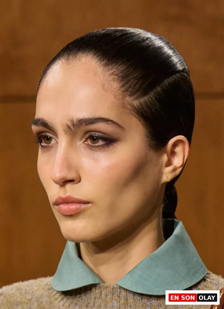 Modelin yakın çekim portresinde, Tory Burch Sonbahar 2026 Ready-to-Wear defilesinden, doğal kalın kaşları, belirgin eyeliner ve parlak nude dudak makyajı dikkat çekiyor; saçları parlak ve arkaya taranmış, kulak arkasında zarif bir örgüyle toplanmış, üzerinde sade örgü bir kazak ve mint yeşili yuvarlak yakalı bir gömlek görülüyor.