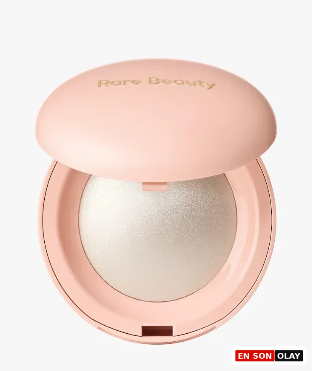 Açık pembe renkli, üst kapağında altın rengi Rare Beauty yazısı yer alan ve içinde ışıltılı, gümüş tonlarında Positive Light Silky Touch aydınlatıcının göründüğü, hafifçe aralanmış kompakt bir makyaj ürününün yakından görünümü.