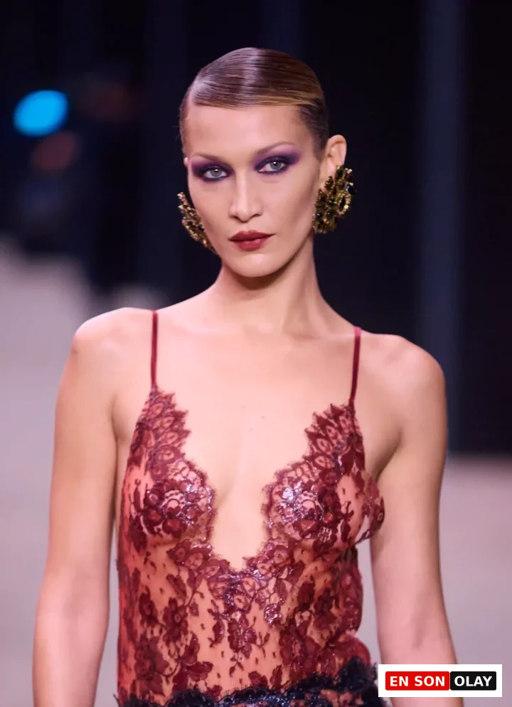 Bella Hadid, Saint Laurent podyumunda mor tonlarında iddialı göz makyajı ve bordo rujuyla, ıslak görünümlü geriye taranmış saçları ve gösterişli altın rengi küpeleriyle zarif bir şekilde yürürken, derin yakalı kırmızı dantel bluzuyla dikkat çekiyor.