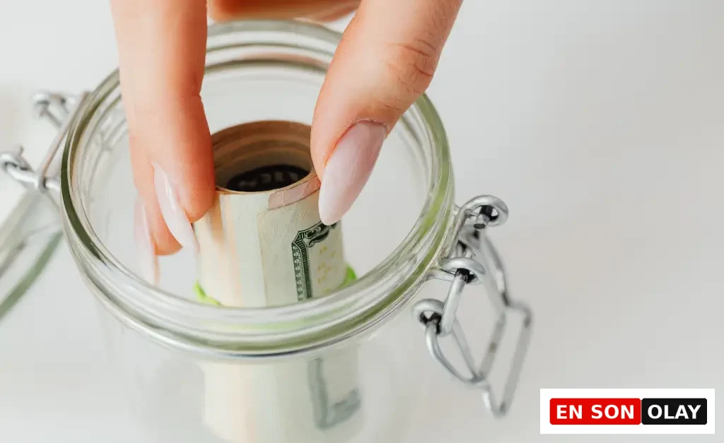 Bakımlı tırnaklara sahip bir elin, rulo yapılmış dolar banknotlarını şeffaf bir cam kavanozun içine dikkatlice yerleştirmesi, tasarruf etmenin ve reel faiz gibi ekonomik göstergelerin birikimler üzerindeki etkisinin önemini akla getiriyor.