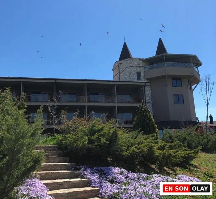 Parlak mavi gökyüzü altında, mor çiçeklerle kaplı taş bir merdivenin çevrelediği yemyeşil çalılıklar ve ağaçların arasından yükselen, kale benzeri kuleleri ve katlı balkonları olan etkileyici bir otel veya tesis binası görülüyor, gökyüzünde kuşlar uçuşuyor.