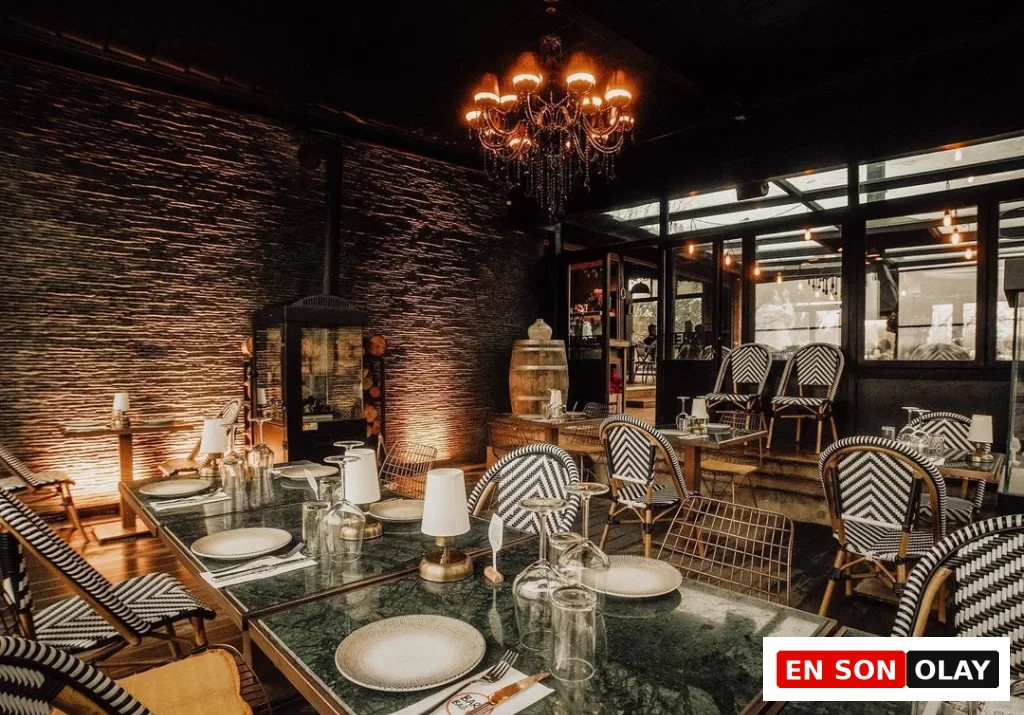 Loş ışıklı, şık bir restoran veya barın iç mekanında, koyu yeşil mermer üst yüzeyli masalar ve belirgin siyah beyaz zikzak desenli dokuma sandalyeler, arka plandaki dokulu koyu taş duvar, odunluklu modern bir şömine ve tavandan sarkan işlemeli zarif avize ile sıcak ve davetkar bir ambiyans hakim.