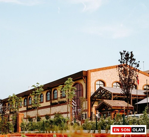 Tuğla renkli cephesi, çeşitli kemerli ve dikdörtgen pencereleri, ön kısmında belirgin bir yüksek kemerli girişi veya penceresi bulunan, modern ve geleneksel unsurları harmanlayan büyük bir bina, çevresi yeşil ağaçlar ve bitkilerle çevrili, aydınlık bir günde gökyüzünün altında göz alıcı bir şekilde yükseliyor.