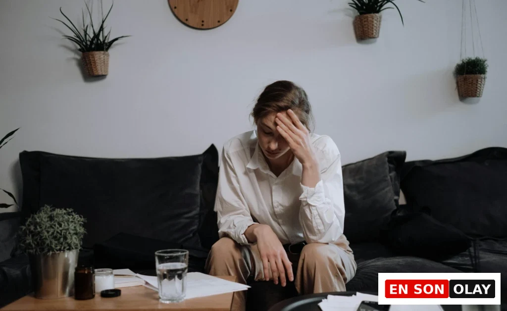 Koyu gri bir koltukta, beyaz gömlek ve bej pantolon giymiş, elini alnına dayayarak öne eğilmiş, bitkin ve düşünceli görünen genç bir kadın, ev ortamında tükenmişlik sendromu yaşadığını düşündüren bir hali yansıtıyor; önündeki ahşap sehpada bir bardak su, dağınık kağıtlar ve birkaç küçük bitki göze çarpıyor.
