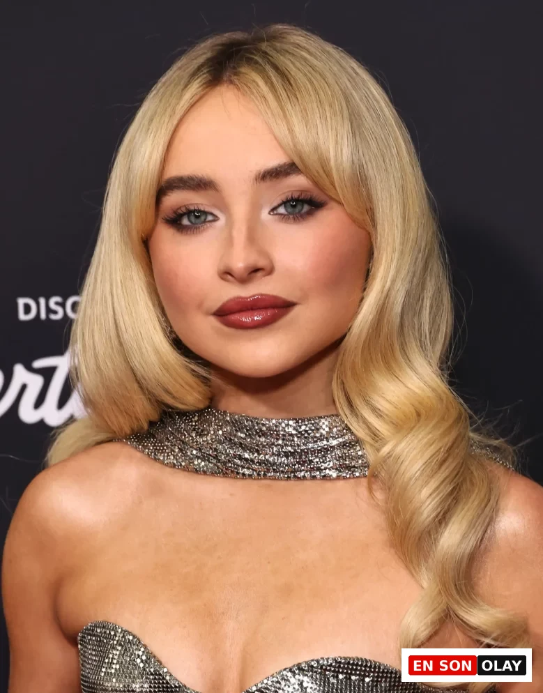 Sabrina Carpenter'ın omuzlarına dökülen sarı, dalgalı saçları ve yüzünü çevreleyen modern perçem modeli ile göz kamaştırıcı makyajının vurgulandığı, gümüş rengi parıltılı straplez elbisesi içinde çekilmiş yakın plan bir portresi.