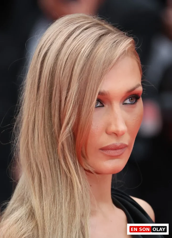 Bella Hadid'in açık kumral, uzun ve yandan ayrılmış saçlarının sol omzuna döküldüğü, kırmızı tonlarında göz makyajı ve doğal dudaklarıyla yukarı doğru baktığı bu yakın plan görüntü, modern perçem modellerine ilham veren zarif bir stil sunuyor.