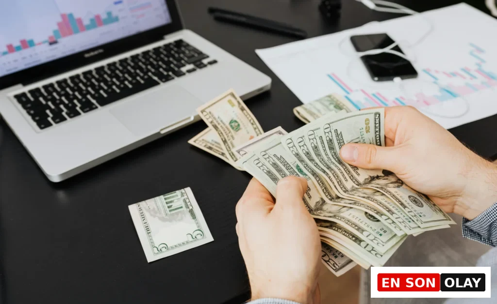 Bir kişi, masa üstünde laptop ve grafikler varken elindeki Amerikan dolarlarını sayıyor.