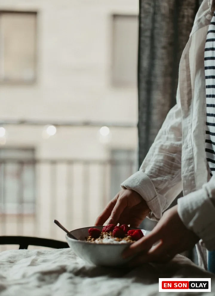 Açıklamalı bir mindful beslenme örneği olarak, keten örtülü bir masaya taze frambuaz ve granolayla süslenmiş yoğurt kasesini özenle yerleştiren bir kişinin elleri, yemeğe başlamadan önce yavaşlamanın ve duyuları devreye sokmanın önemini vurguluyor.