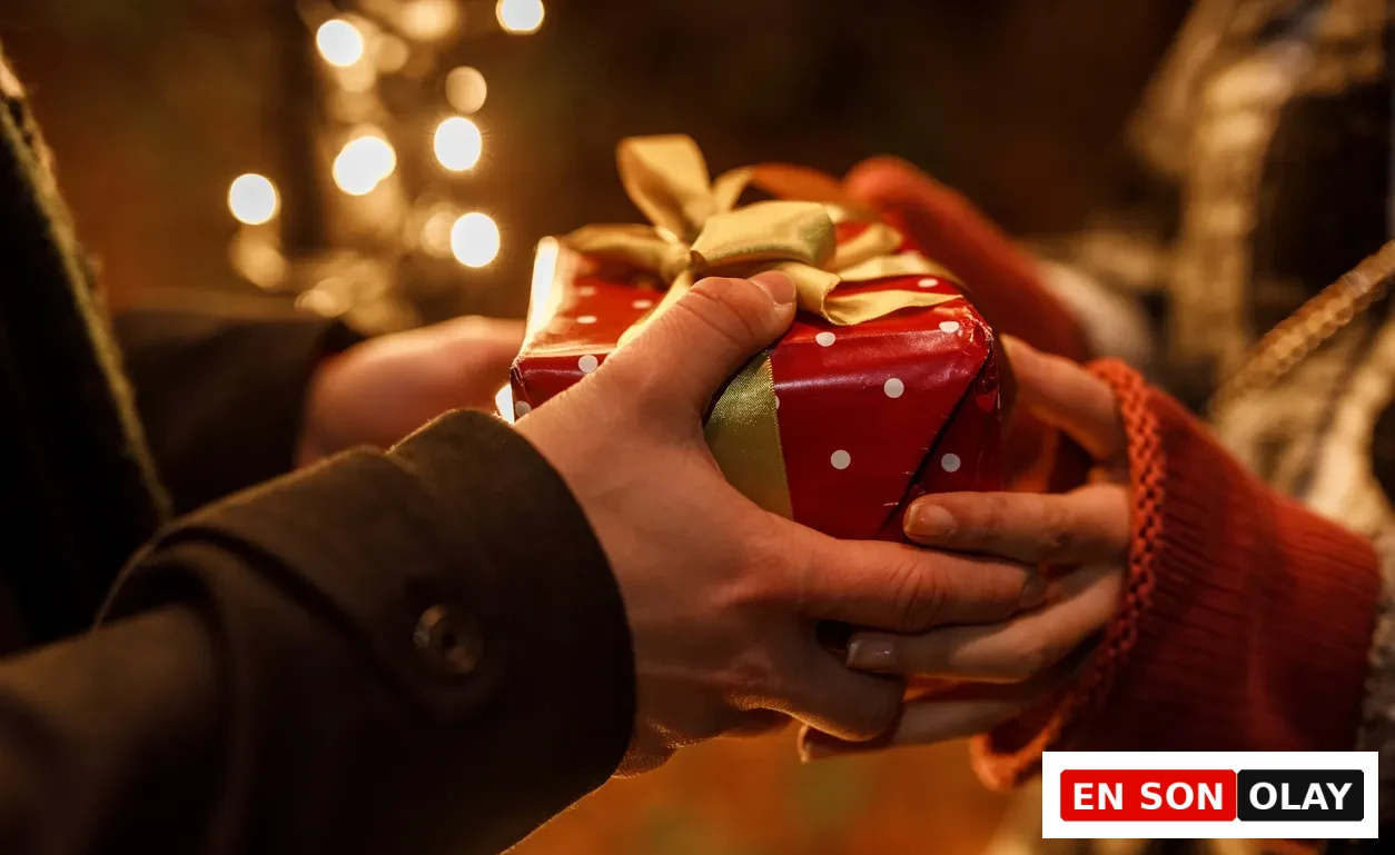 Karanlık bir paltonun kolu ve kırmızı örgü bir kazağın manşeti görünen iki el, arka planda sıcak bokeh ışıklarıyla aydınlanan romantik bir ortamda, altın kurdeleli kırmızı puantiyeli bir hediye kutusunu hassasiyetle değiş tokuş ediyor.
