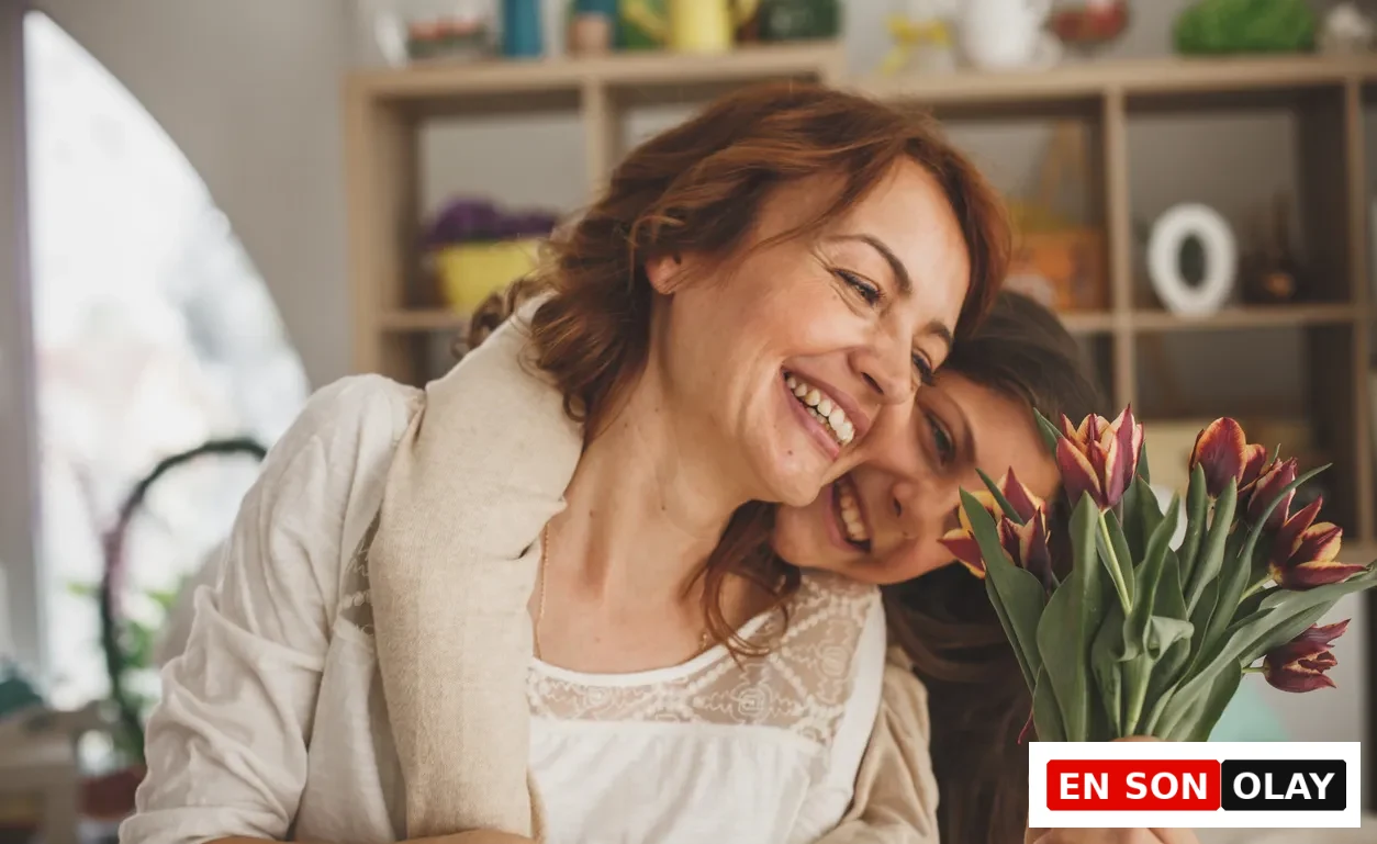Gülümseyen, mutlu bir anne ve ona sarılarak başını omzuna yaslamış genç kızı, kızının elinde tuttuğu canlı kırmızı ve sarı tonlarında lale buketiyle neşeli ve sıcak bir anı paylaşıyorlar.