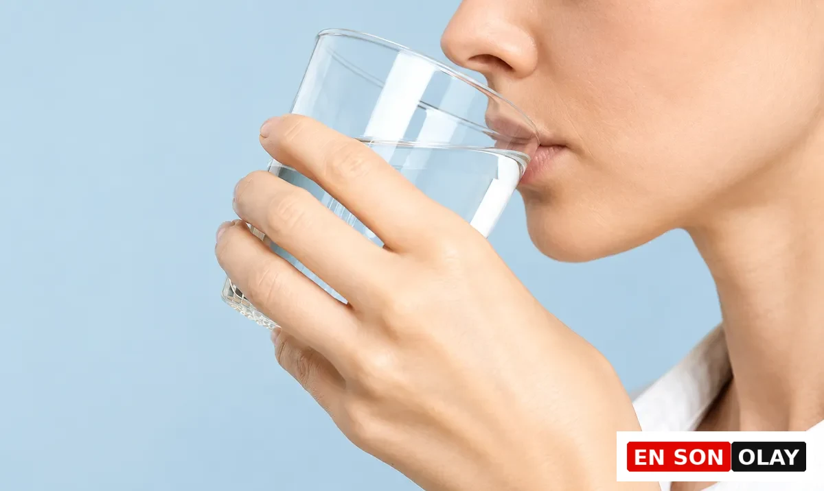 Açık mavi bir arka planın önünde, beyaz yakalı bir gömlek giyen bir kişinin elinde tuttuğu şeffaf bardaktan ağzına su akıttığı net bir yakın çekim, özellikle besin zehirlenmesi gibi durumlarda vücudun hidrasyonu için düzenli su tüketiminin ne kadar hayati olduğunu gösteriyor.