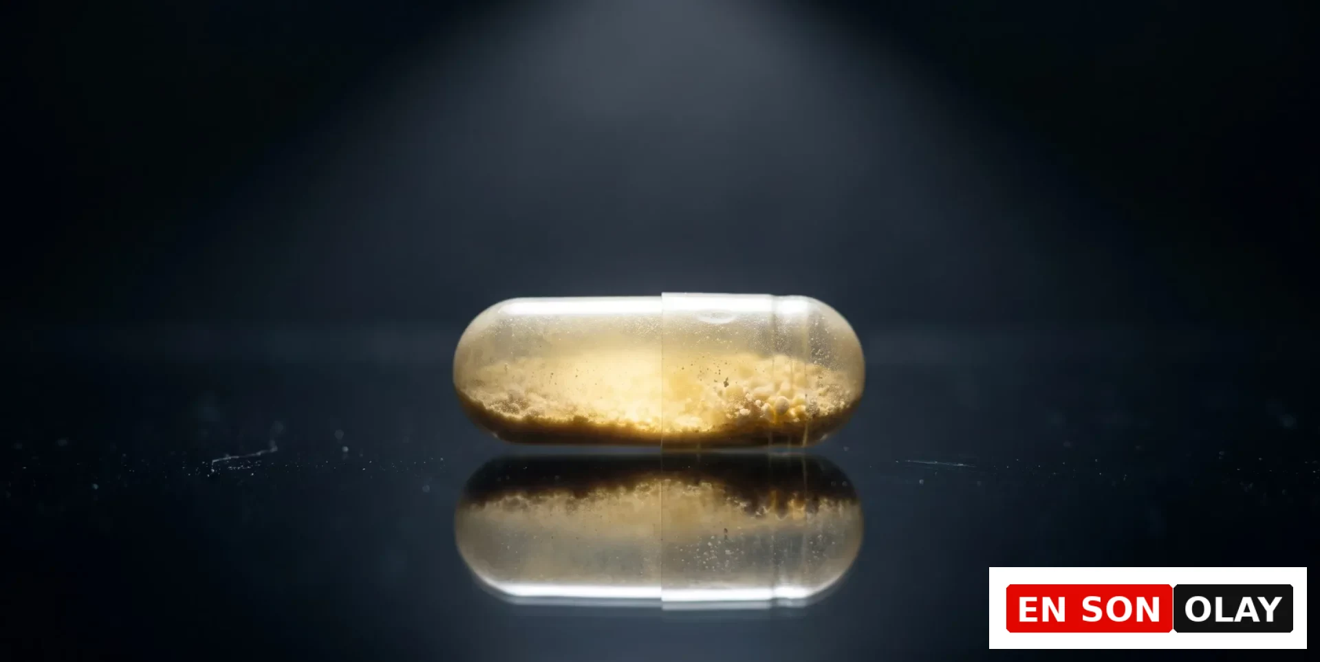Bir spot ışığıyla yukarıdan aydınlatılmış, içinde sarımsı kahverengi granül tozlar bulunan şeffaf bir B6 vitamini kapsülü, yansıtıcı koyu renkli bir yüzeyde yatay olarak duruyor ve yüzeydeki toz partikülleri ışıkla vurgulanıyor.