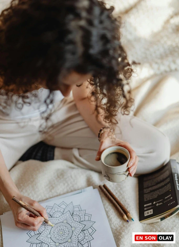 Bir kadın, battaniyesine sarınmış şekilde mandala boyarken bir yandan kahve içiyor; sanatla mindfulness anı.